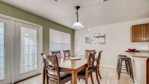 Un comedor con una mesa de madera, seis sillas, una lámpara en el techo y una cocina con taburetes y armarios.