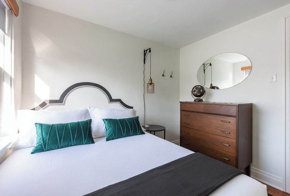 A neatly arranged bedroom with a bed, a dresser, a round mirror, and a hanging light fixture.