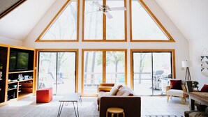A spacious living room with a high ceiling, large windows, a ceiling fan, and a television.