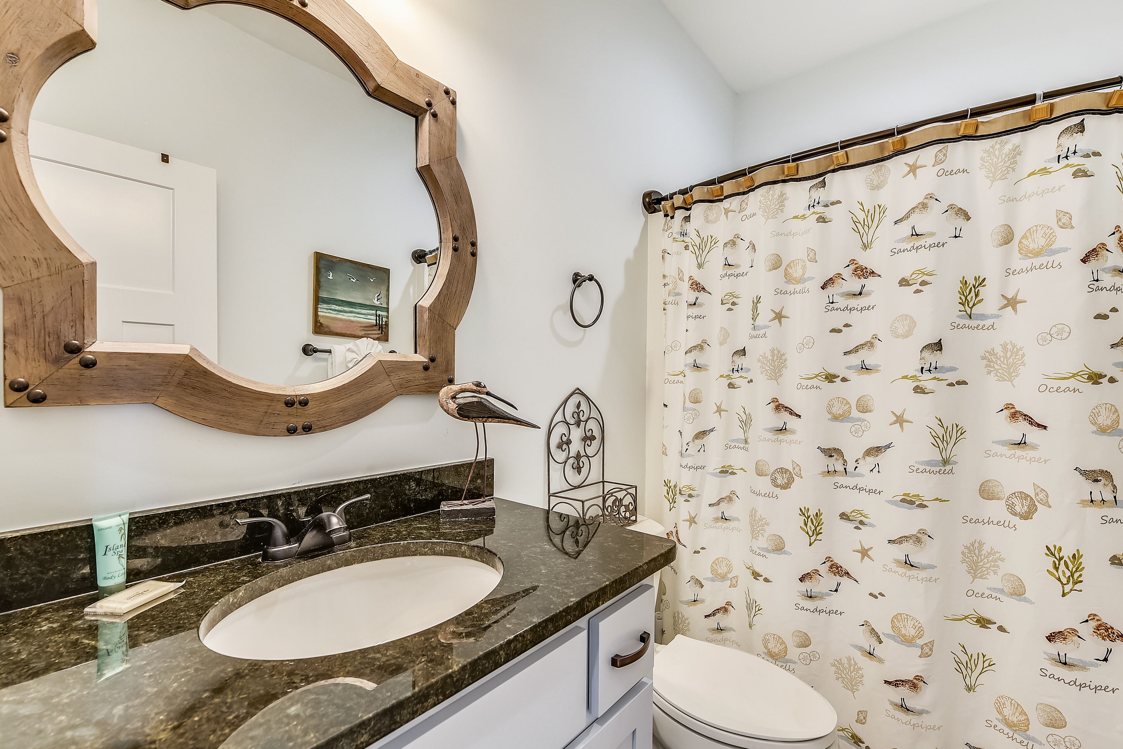 Un bagno con uno specchio a cornice di legno, un lavabo con piano in marmo, un water e una tenda da doccia con motivo marino.