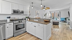 Une cuisine moderne avec des meubles blancs, des comptoirs en granit, des appareils électroménagers en acier inoxydable, des luminaires suspendus et un ventilateur de plafond.