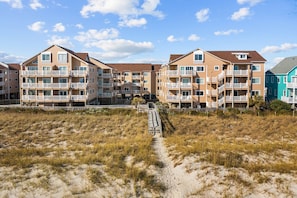 A beachfront residential building with multiple balconies, a sandy path leading to the entrance, and a clear sky. - "Cast-a-waves" Make every day a Holiday! (Carolina Beach)