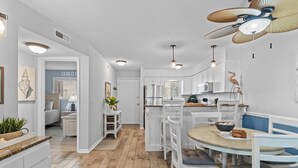 Une cuisine avec des meubles blancs, une table à manger avec des chaises, un ventilateur de plafond et une vue sur la chambre adjacente.