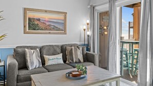 Un salon chaleureux avec un canapé, une table basse agrémentée d’objets décoratifs, un tableau représentant un paysage encadré et une vue sur l’extérieur.