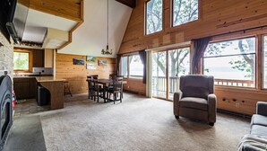 A spacious living area with a high ceiling, wooden walls, a dining table, and a large window offering a view of the outdoors. -  Riverview Cabin home! (Stoddard)