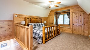 A cozy bedroom with a wooden headboard, a bed with a plaid bedspread, a wooden railing, a ceiling fan, and a window with curtains. -  Riverview Cabin home! (Stoddard)