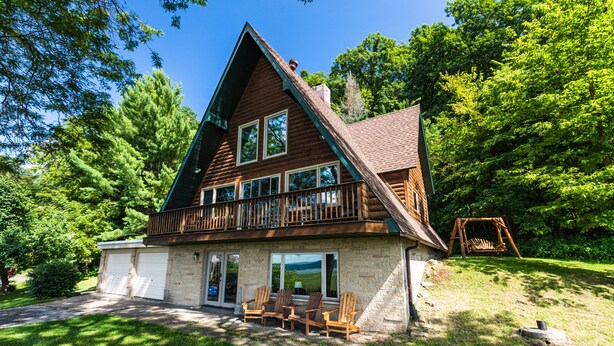 A log cabin with a balcony, two garage doors, and a patio with four Adirondack chairs. -  Riverview Cabin home! (Stoddard)