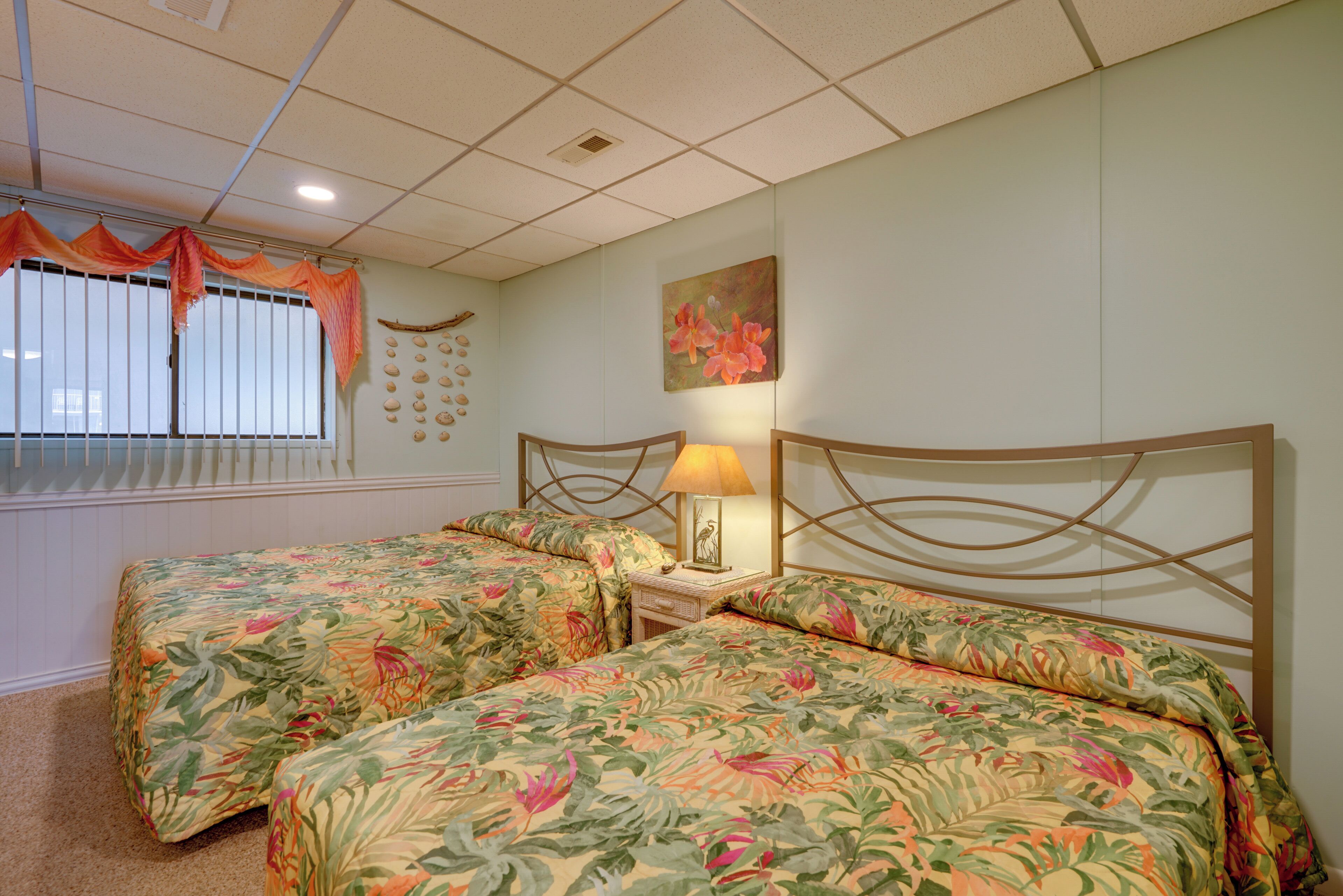 A hotel room with two beds, a window with blinds, a wall-mounted artwork, and a nightstand with a lamp.
