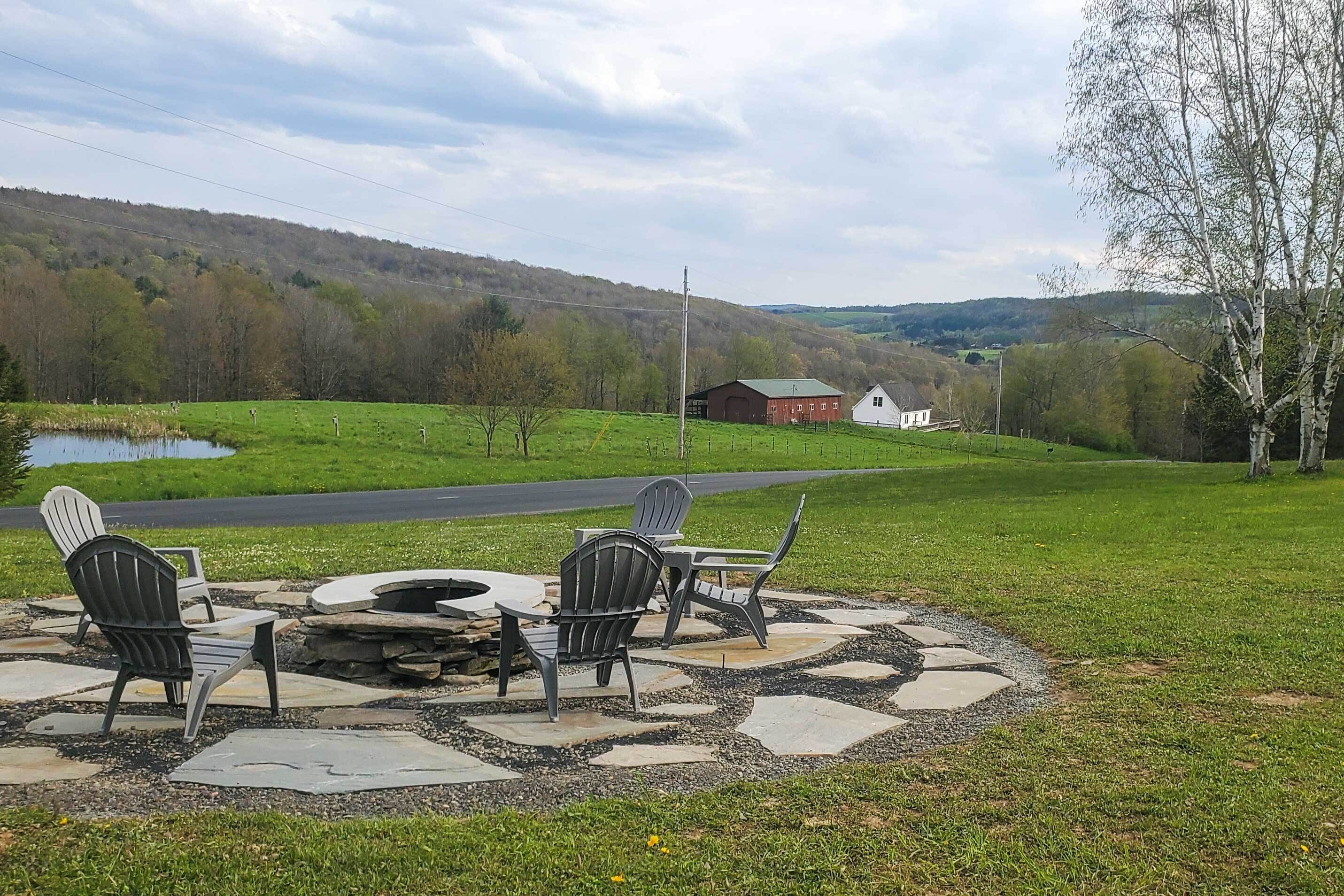 En terrasse med Adirondack-stole og bålsted, med udsigt over et grønt landskab med en sø og en vej.