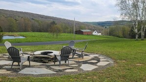 Eine Terrasse mit Adirondack-Stühlen und einem Feuerkorb, mit Blick auf eine grüne Landschaft mit Teich und einer Straße.