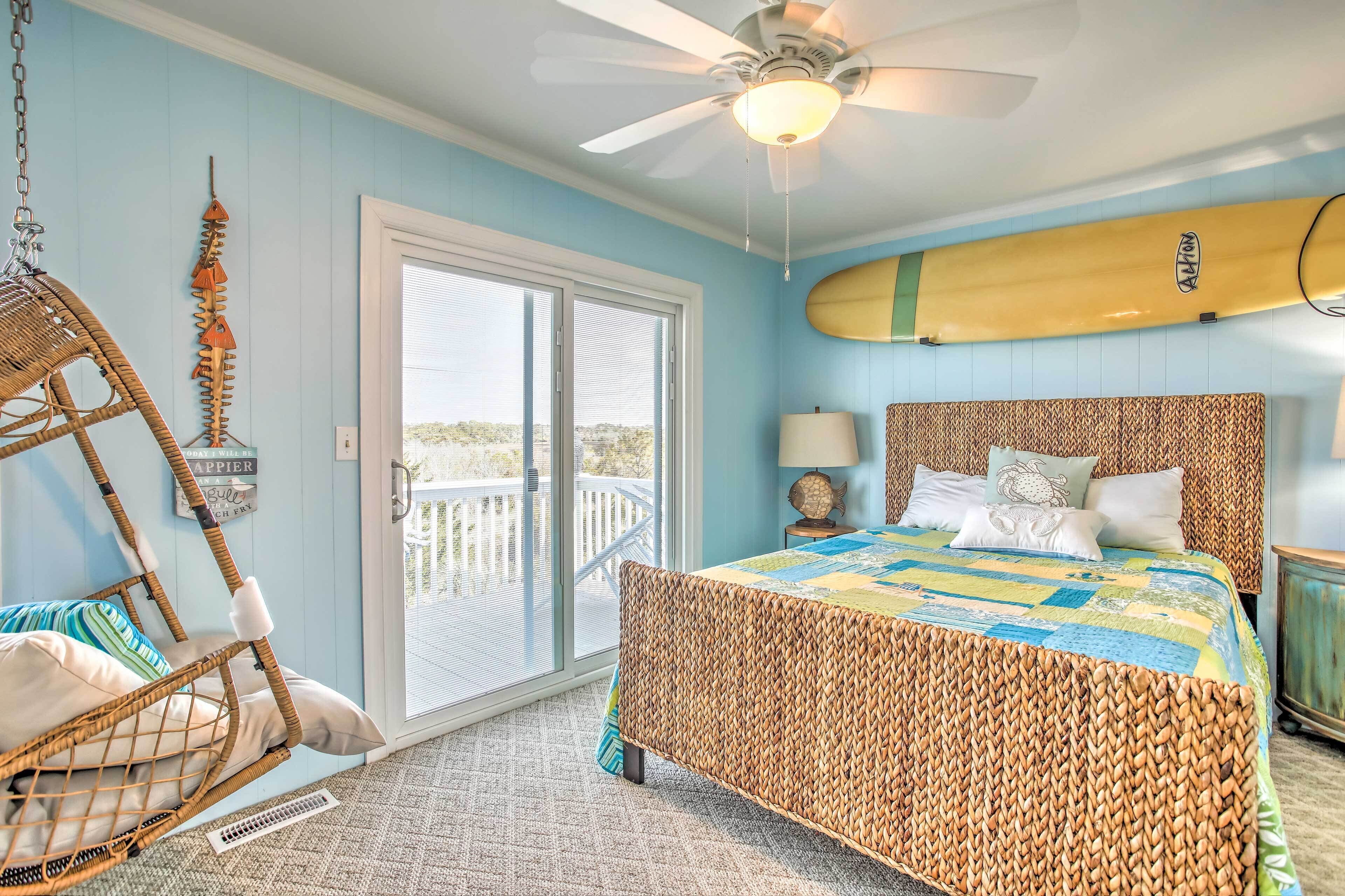 A bedroom with a wicker bed, a ceiling fan, a surfboard on the wall, and a balcony with a view.