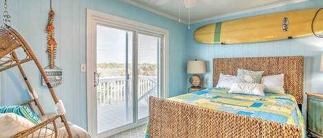 A bedroom with a wicker bed, a ceiling fan, a surfboard on the wall, and a balcony with a view.