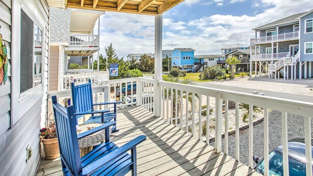 Una terraza de madera con sillas azules y una mesa, con vista a una playa y casas al fondo.