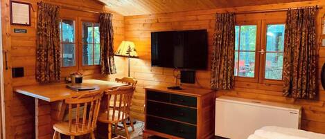 A log cabin room with a wooden desk, two chairs, a television, a radiator, and a bed.