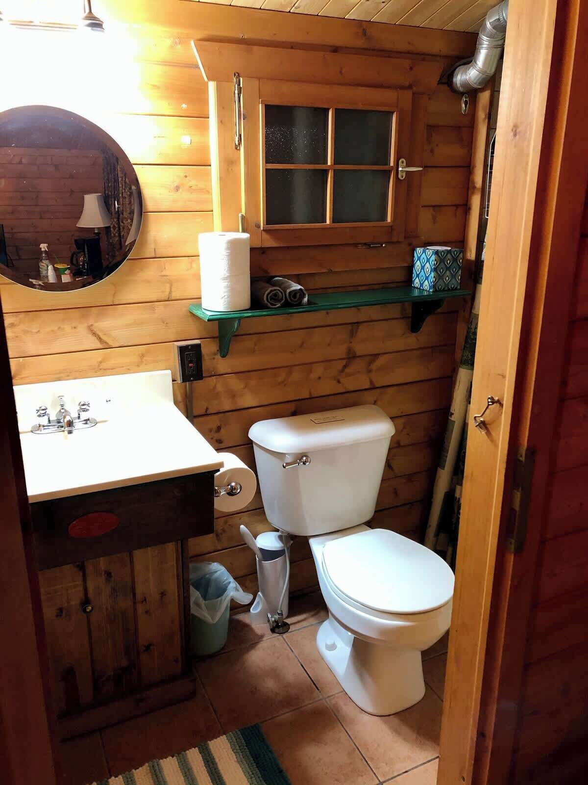 Une petite salle de bain en bois, aménagée dans une cabane, avec un lavabo, des toilettes et une fenêtre située au-dessus des toilettes.