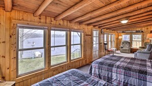 A cabin with wooden walls and ceiling, two beds with plaid bedding, a chair, a table, and large windows overlooking a lake. - Lakefront 1930’s Log Cabin w/ Dock & Fire Pit! (Rice Lake)