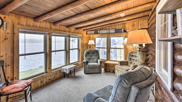 A cozy cabin interior with wooden walls and ceilings, large windows overlooking a body of water, and comfortable seating including a chair and a sofa. - Lakefront 1930’s Log Cabin w/ Dock & Fire Pit! (Rice Lake)