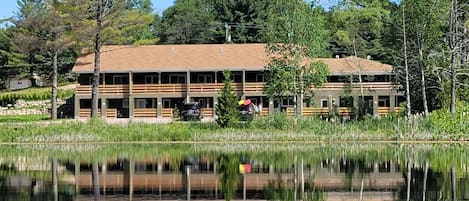 A two-story building with a flat roof, surrounded by trees and a body of water.