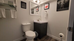 Ein modernes Badezimmer mit Toilette, Waschbecken und Spiegel. An der Wand befinden sich Bilder in Rahmen, ein Handtuchhalter und ein Mülleimer.