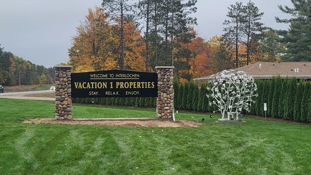 Ein Willkommensschild für Vacation 1 Properties mit einer Säule aus Stein, vor dem Hintergrund von Herbstbäumen und einem gepflegten Rasen.