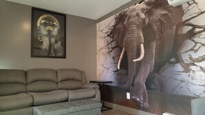 Ein Wohnzimmer mit einem grauen Sofa, einem gerahmten Kunstwerk, das ein Frauenporträt zeigt, und einem großen Wandgemälde mit einem Elefanten.
