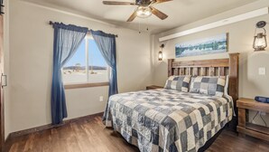 A bedroom with a wooden bed, a ceiling fan, a window with curtains, a nightstand, and a wall-mounted lamp. - Family-Friendly Old West Cabin Near YNP (Island Park)