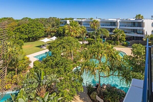 A resort with a pool surrounded by lush greenery and palm trees, modern buildings in the background, and a clear blue sky.