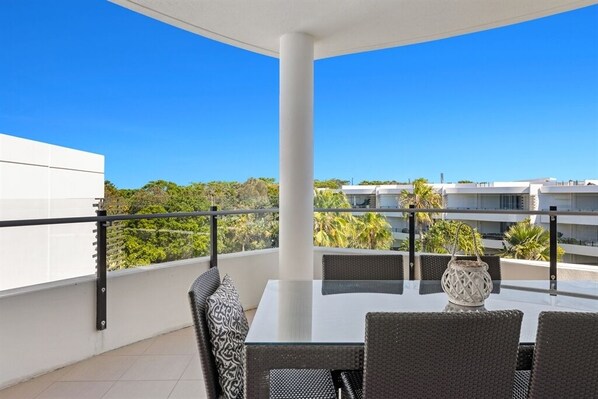 A balcony with a glass table and wicker chairs, overlooking a residential area with palm trees and buildings.