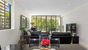 A modern living room with a glass table centerpiece, black leather sofas, and a large sliding glass door leading to a balcony with greenery.