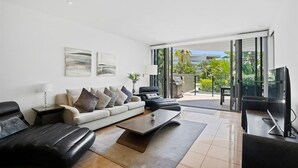 A modern living room with a sofa, coffee table, and TV. There is a balcony with a view of greenery and a building.