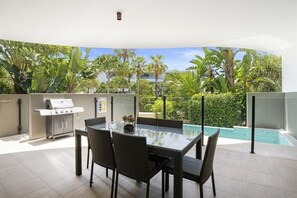 A modern outdoor dining area with a glass table and wicker chairs, a barbecue grill, and a pool in the background.