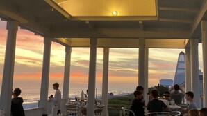 Een overdekt terras met uitzicht op de oceaan en een stadsachtergrond tijdens de zonsondergang.