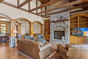 A spacious living room with a stone fireplace, leather sofas, wooden bookshelves, and a dining area with a chandelier.