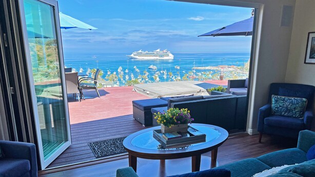 A balcony with a view of the ocean and a cruise ship, furnished with a sofa, chairs, and a table with a flower arrangement.