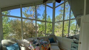 Uma sala de sol com grandes janelas, uma mesa com toalha florida e duas cadeiras com almofadas azuis.