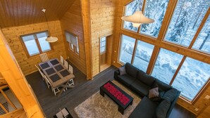 Interior acogedor de una cabaña con comedor, sofá y grandes ventanas que dan a una vista nevada de un bosque.