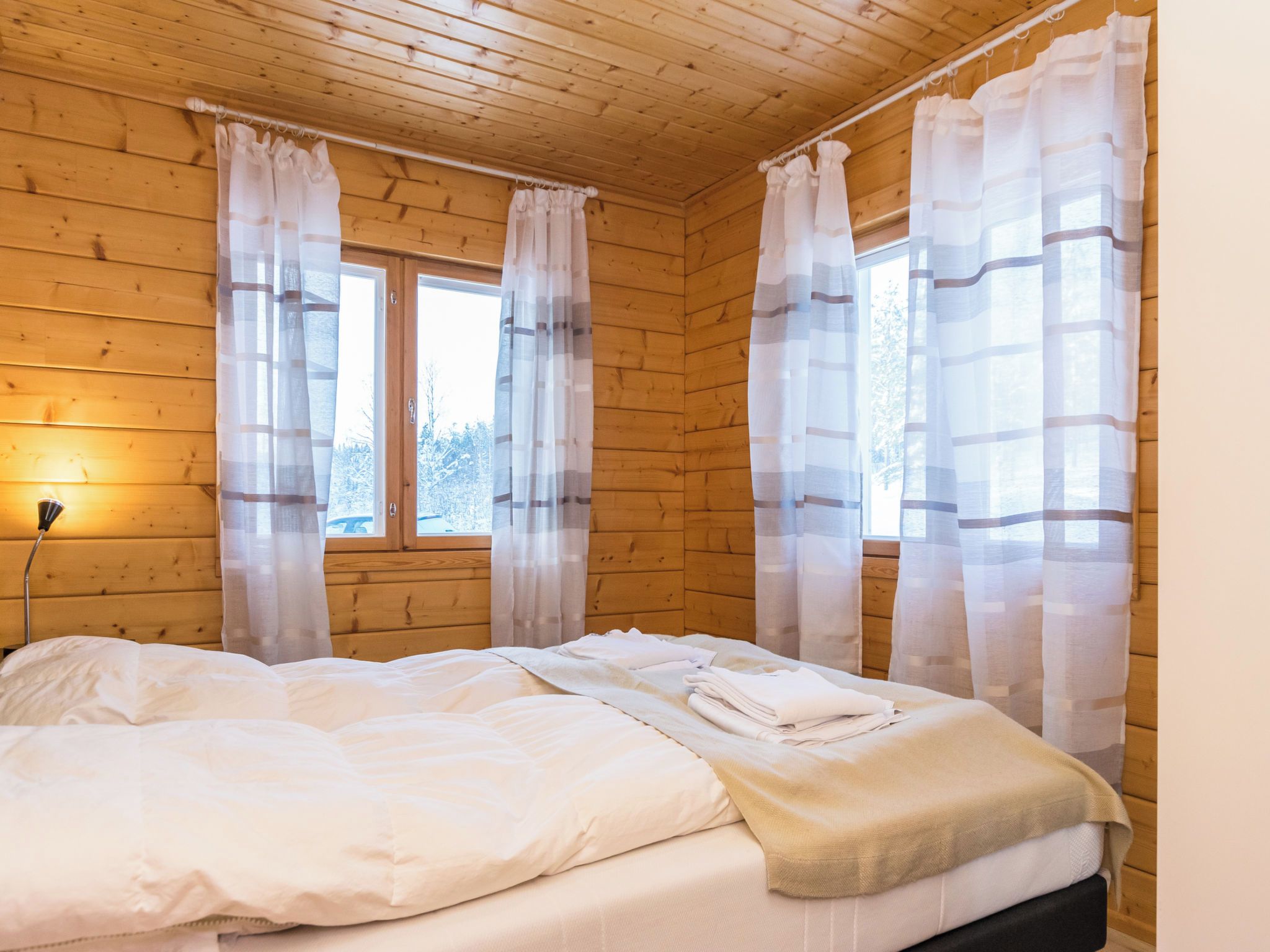 Quarto com paredes de madeira, cama com roupa de cama branca e cortinas leves nas janelas.