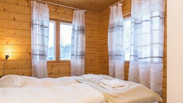 Quarto com paredes de madeira, cama com roupa de cama branca e cortinas leves nas janelas.