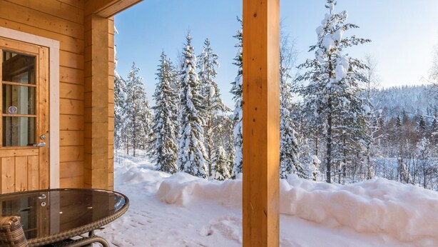 Una cabaña de madera con puerta de vidrio y una mesa con sillas afuera, con vista a un bosque nevado.
