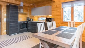 Una cabaña moderna de troncos con cocina que presenta armarios oscuros, horno integrado y microondas. Hay un comedor con mesa de madera y sillas blancas.