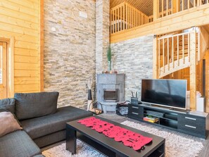 A cozy living room with a stone wall, a fireplace, a television, and a staircase. - Villa oka by Interhome (Hyrynsalmi)