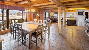 A spacious dining area with a large wooden table and chairs, a cozy seating area with a sofa and a fireplace, and a view of the surrounding landscape through large windows. - Summer Retreat, Dog Friendly, Fenced Yard, Hot Tub, Game Room, Fire Pit (Flagstaff)