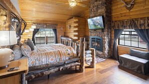 A log cabin interior with a bed, a stone fireplace, a television, and a ceiling fan. - Summer Retreat, Dog Friendly, Fenced Yard, Hot Tub, Game Room, Fire Pit (Flagstaff)