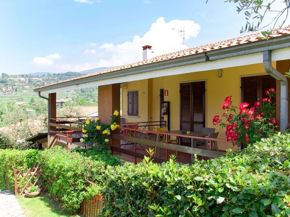 A two-story house with a balcony, a garden with various plants, and a view of the countryside.