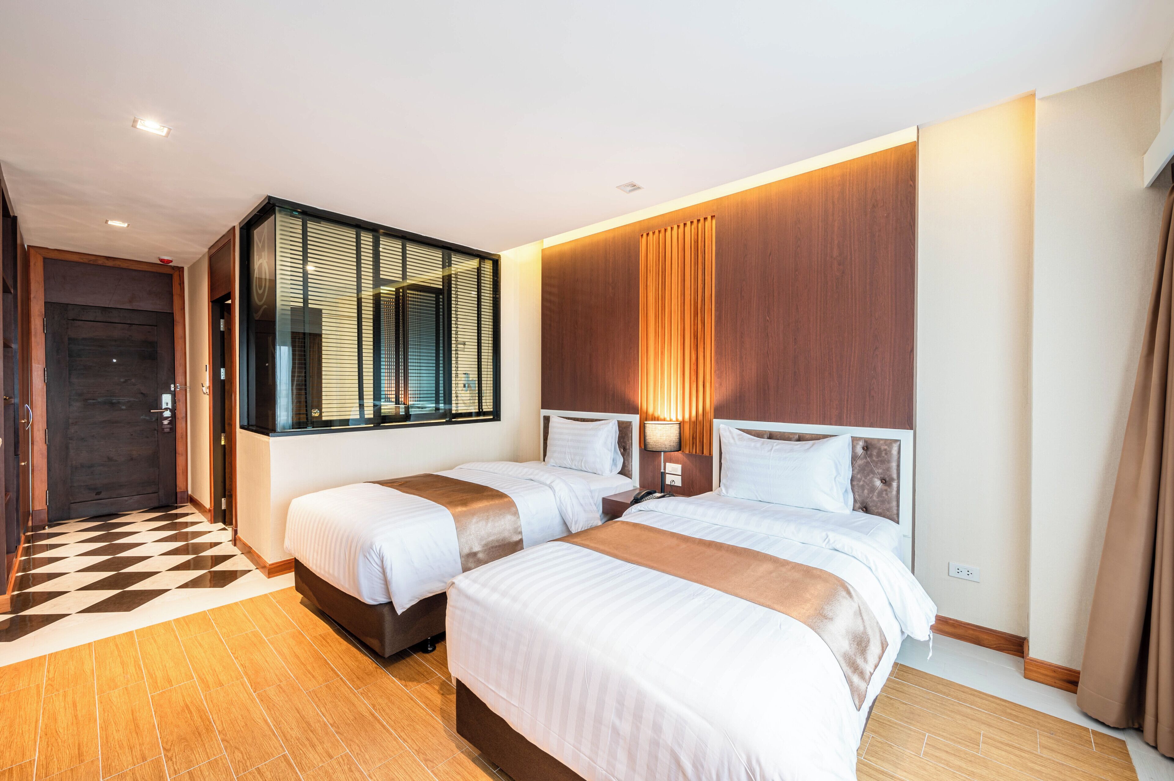 A hotel room with two beds, a checkered floor, a wooden wall, and a large window with blinds.