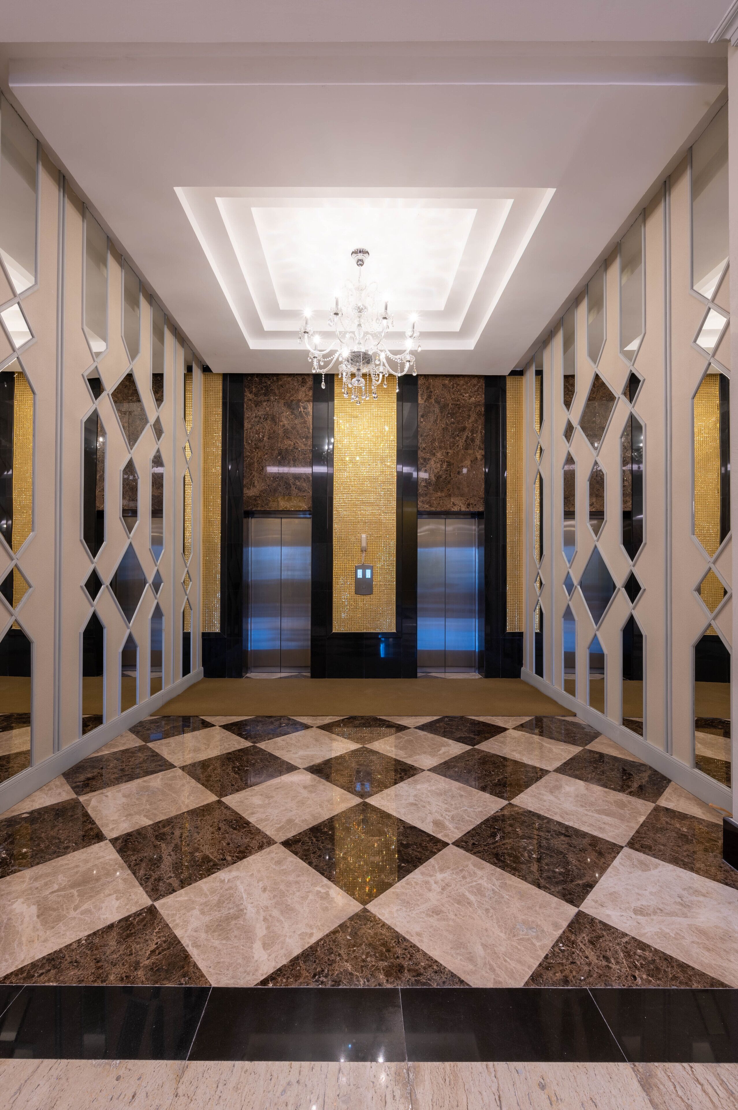 A spacious lobby with a checkered marble floor, a chandelier, elevators, and a decorative wall with geometric patterns.