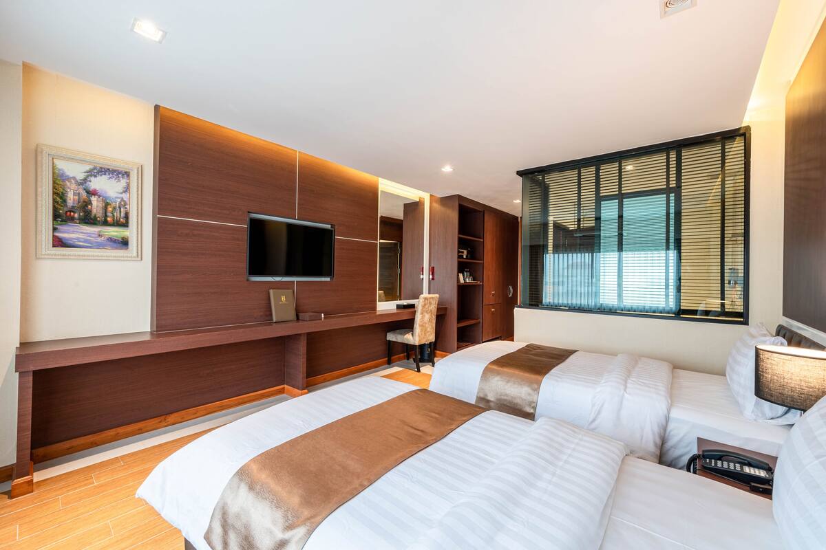 A modern hotel room with two beds, a TV, a desk, and a large window with blinds.