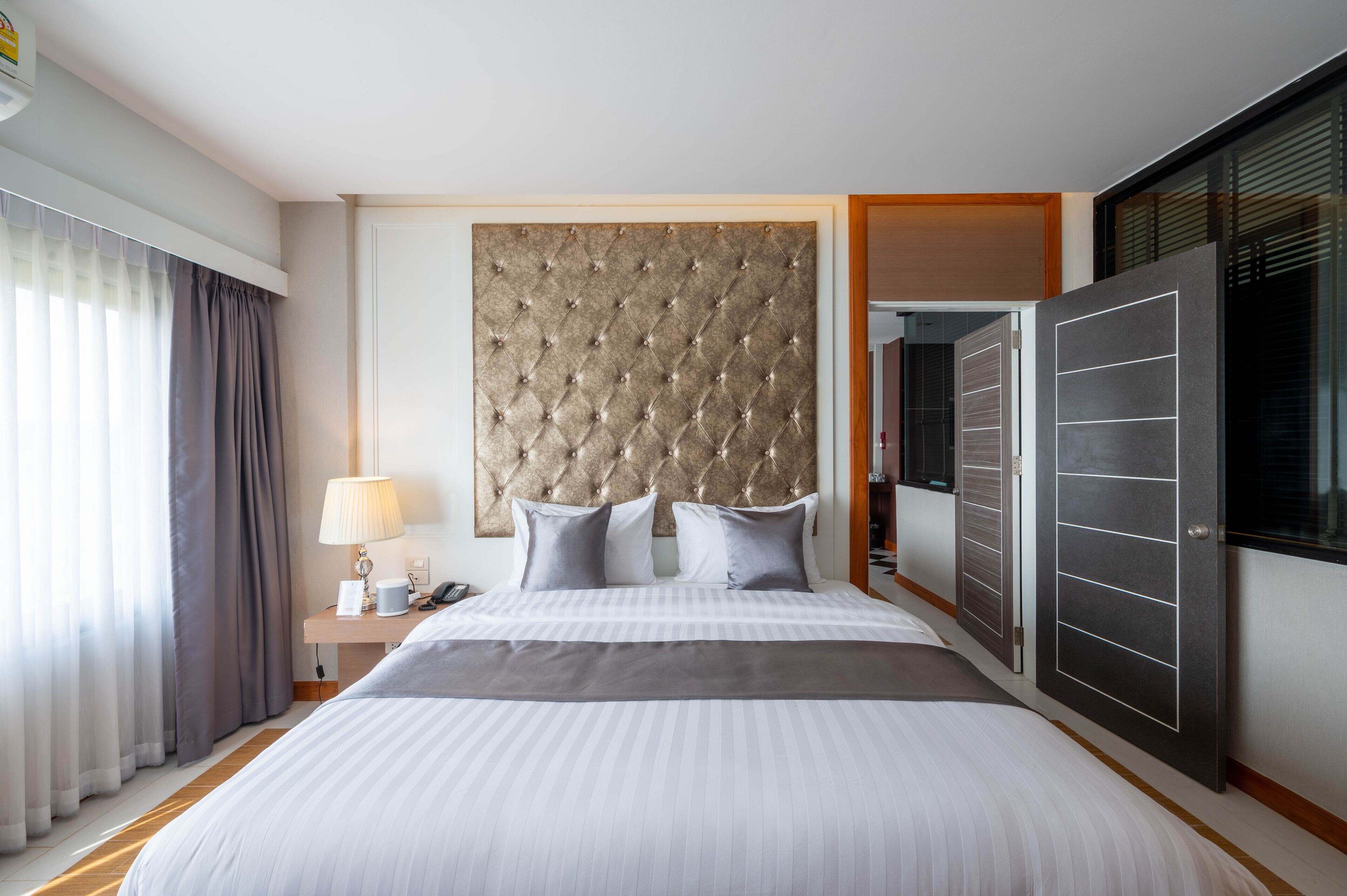 A hotel room with a large bed, a tufted headboard, a bedside table with a lamp, and a wardrobe.