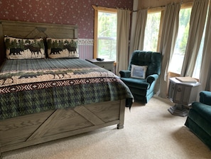 A bedroom with a large bed, a green armchair, a small side table, and a window with curtains. - Kenai River Frontage - Bings Landing  (Sterling)