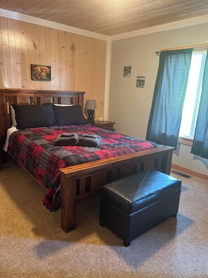 Un dormitorio con cabecera de madera, colcha acolchada, mesita de noche con lámpara y ventana con cortinas.